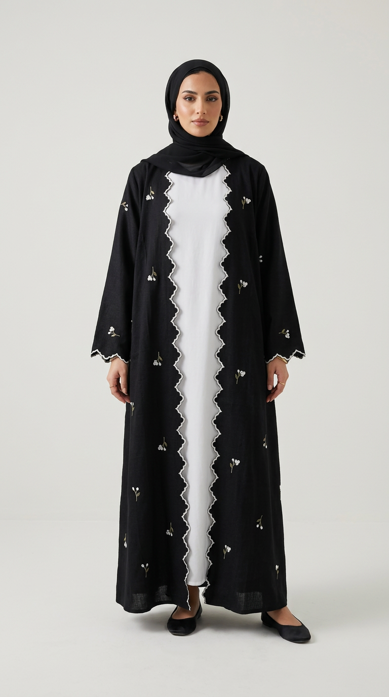 A woman modeling a long black abaya with delicate white floral embroidery and a scalloped white inner lining, worn over a white dress with a black hijab, on a light gray background.
