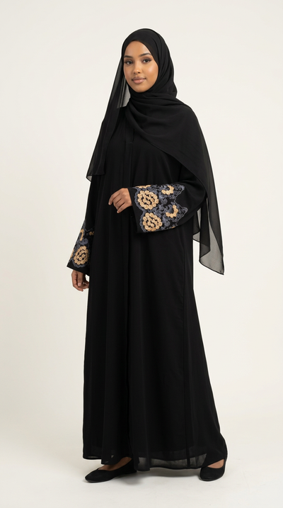 A woman modeling a black abaya with long sheer sleeves featuring intricate gold and beige floral embroidery, paired with a matching black hijab, against a plain light background.