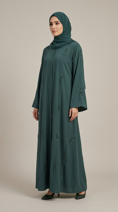 A woman wearing a long-sleeved, floor-length teal green abaya with subtle embroidered details and a matching teal green hijab, standing against a solid light beige background.