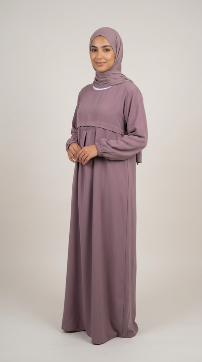 A woman wearing a long-sleeved mauve maxi abaya with a matching hijab, standing against a neutral light gray studio background.