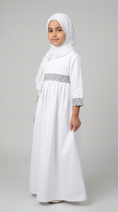 A young girl wearing a white modest dress with a matching hijab, featuring a patterned waistband and sleeve cuffs, standing against a neutral gray studio background.