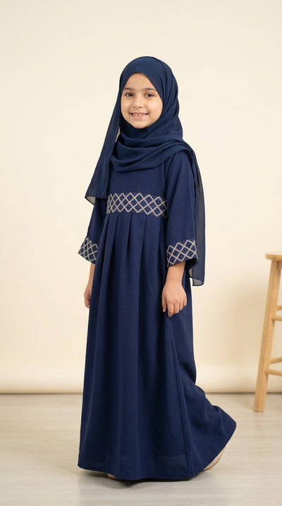 A young girl wearing a navy blue long dress with a matching hijab, featuring white geometric embroidery on the chest and cuffs, standing against a plain beige background.