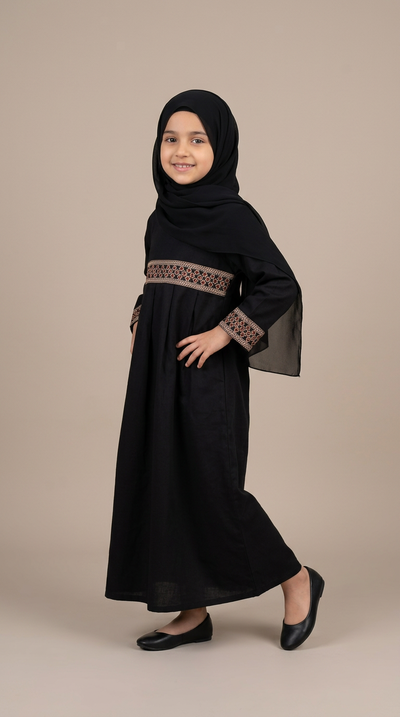 A young girl modeling a black abaya dress with a matching black hijab, featuring brown embroidered details on the waist and cuffs, set against a solid beige studio background.