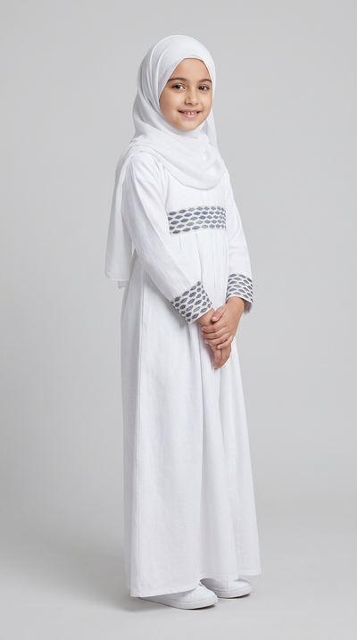 A young girl wearing a white abaya dress with blue patterned trim on the chest and cuffs, paired with a matching white hijab, standing against a light gray studio background.