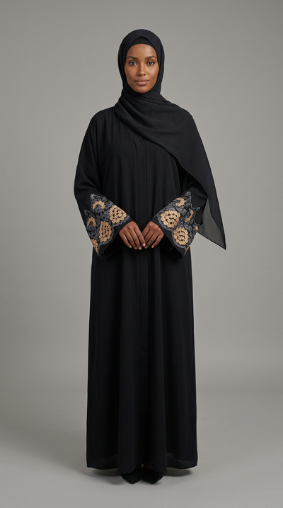 A woman wearing a long black abaya with ornate gold and beige floral embroidery on the sleeves, paired with a matching black hijab, standing against a solid gray studio background.