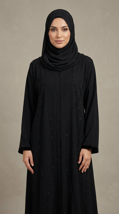 A woman wearing a black abaya with subtle vertical bead embellishments and a matching black hijab, standing against a neutral textured beige studio background.