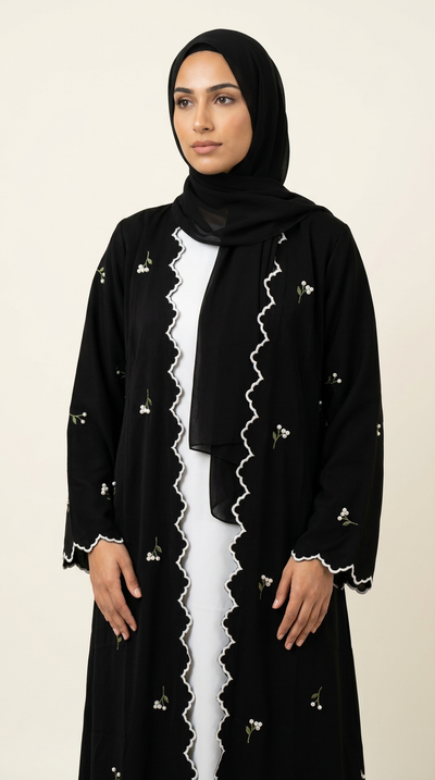 A woman wearing a black abaya with white scalloped trim and small floral embroidery, paired with a matching black hijab and a white inner dress, set against a plain beige background.