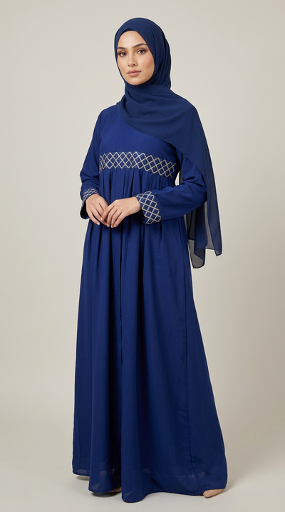 A woman wearing a long royal blue modest dress with a matching hijab and a sheer draped scarf, standing against a plain light beige studio background.