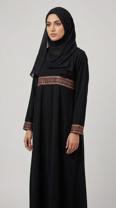 A woman wearing a long black abaya dress with brown embroidered detailing at the waist and cuffs, paired with a matching black hijab, photographed against a plain light gray studio background.