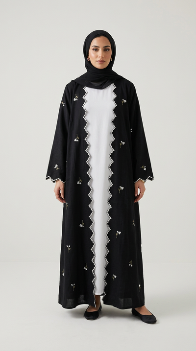 A woman modeling a long black abaya with delicate white floral embroidery and a scalloped white inner lining, worn over a white dress with a black hijab, on a light gray background.