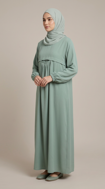 A woman wearing a long-sleeved sage green modest maxi dress with a gathered waist and matching hijab, standing against a solid beige studio background.