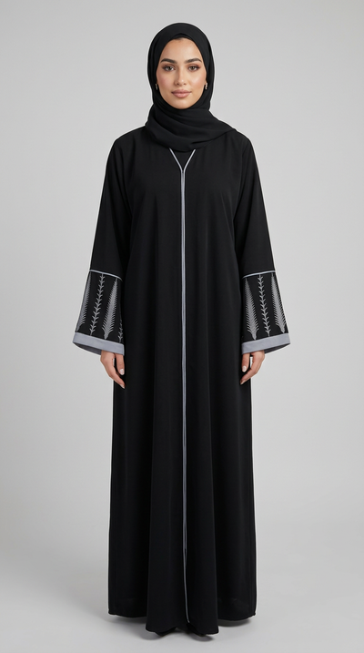 A woman modeling a long black abaya with a front zipper, matching black hijab, and black slip-on shoes against a solid gray studio background.
