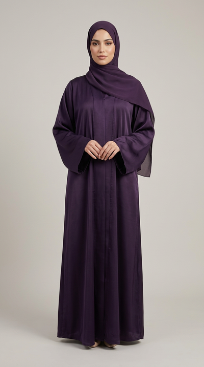 A woman wearing a long, flowing dark purple abaya with a matching headscarf, standing against a neutral light gray studio background.