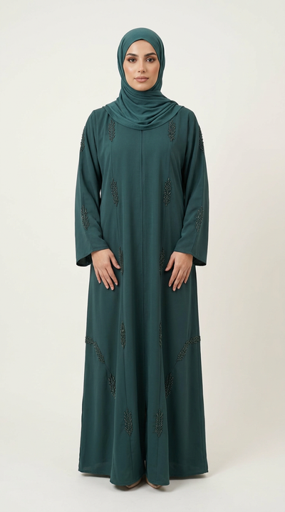 A woman wearing a long-sleeved dark teal green abaya with subtle embroidered details and a matching hijab, standing against a plain light beige studio background.
