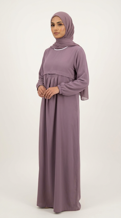 A woman wearing a long-sleeved, floor-length mauve maxi abaya with a matching hijab, standing against a plain off-white studio background.