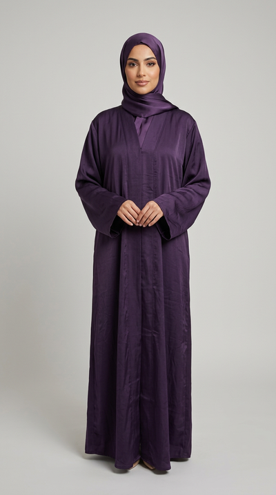 A woman wearing a long, flowing purple abaya and matching hijab stands against a plain light gray studio background.