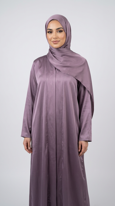 A woman wearing a long, flowing mauve-purple abaya with a matching hijab, standing against a light gray studio background.