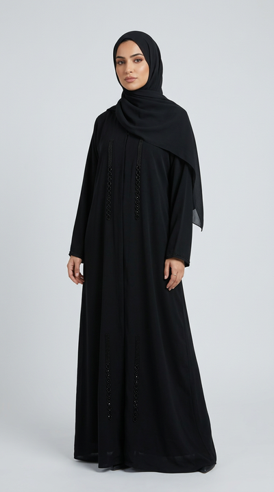 A woman wearing a long black abaya with embroidered details and a matching black hijab, standing against a light gray studio background.