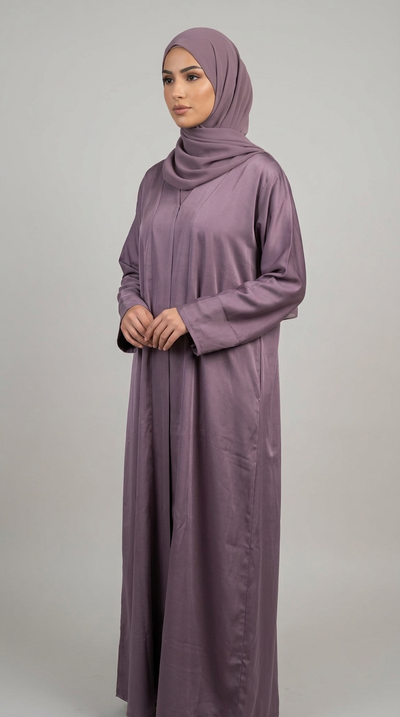 A woman wearing a long, loose-fitting mauve-purple abaya and matching hijab, standing against a plain light gray studio background.