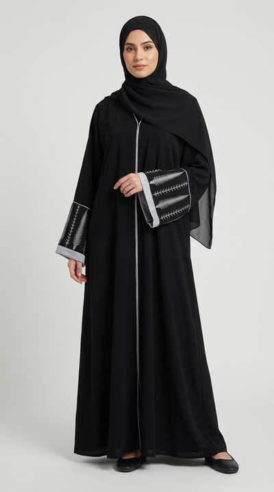 A woman modeling a long black abaya with a front zipper, matching black hijab, and black slip-on shoes against a solid gray studio background.