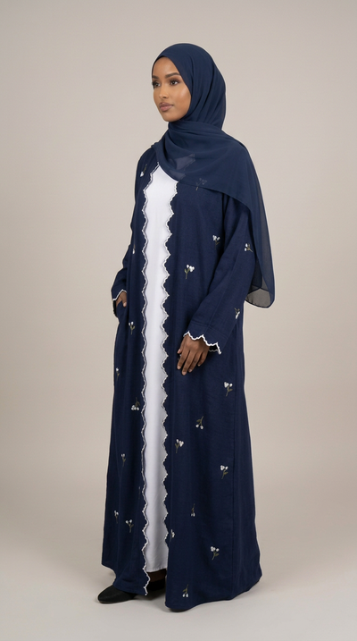 A woman modeling a navy blue long abaya with a white inner dress, matching navy hijab, and small embroidered details, photographed against a solid light beige studio background.