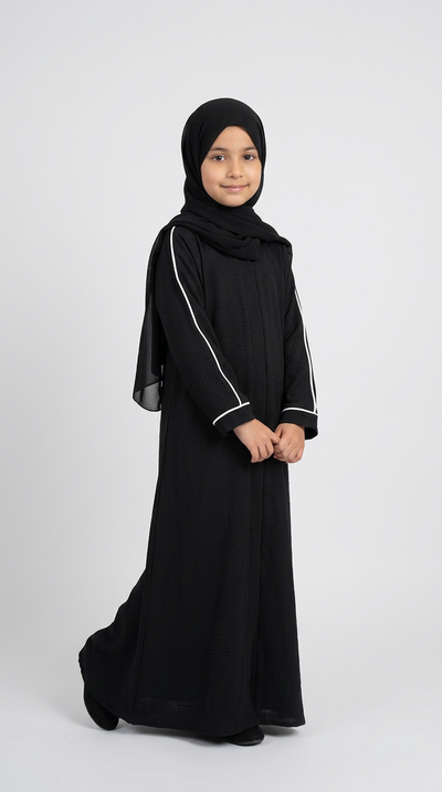 A young girl wearing a black abaya with white piping details, a matching black hijab, and black shoes, standing against a light gray studio background.