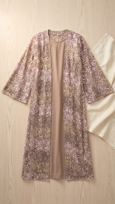 A long-sleeve open-front cardigan or robe with a beige inner lining and an allover floral lace overlay in dusty pink and gold, laid flat on a light wood background next to a cream fabric prop.