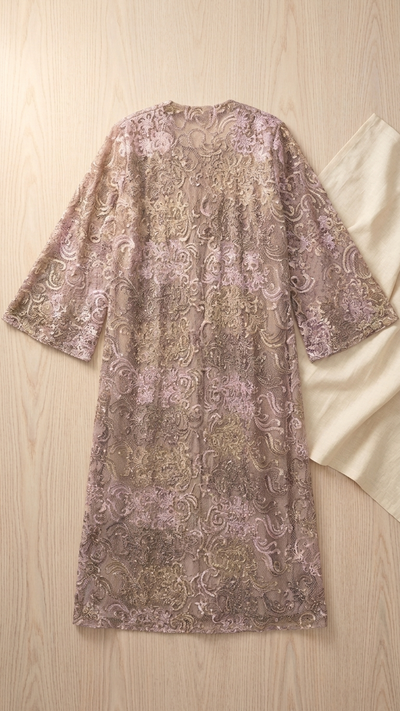 A lavender and beige patterned lace tunic dress with three-quarter sleeves, laid flat on a light wood background next to a cream fabric prop.