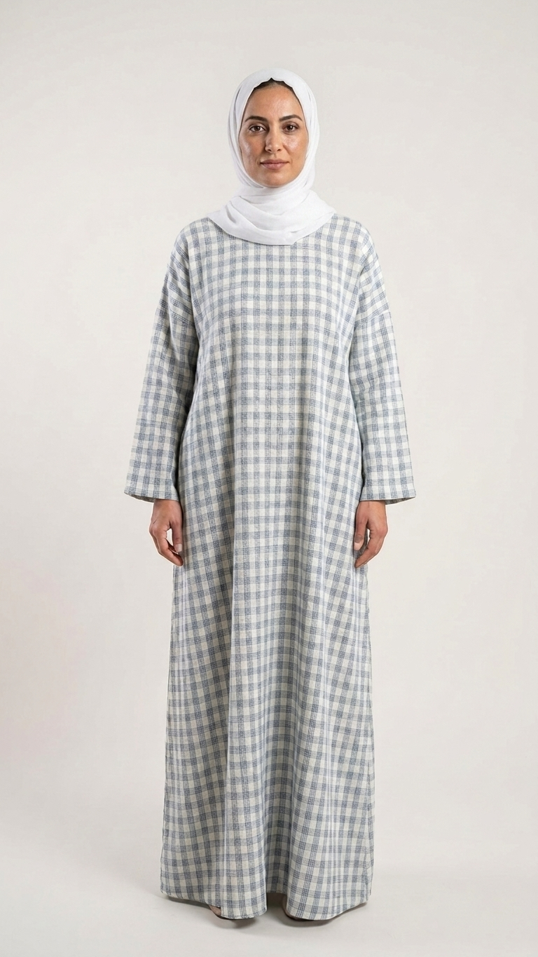 A woman wearing a long-sleeved, floor-length checkered abaya in gray and white, paired with a white hijab, standing against a plain light gray studio background.