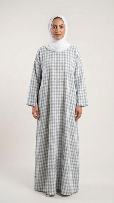 A woman wearing a long-sleeved, floor-length checkered abaya in gray and white, paired with a white hijab, standing against a plain light gray studio background.