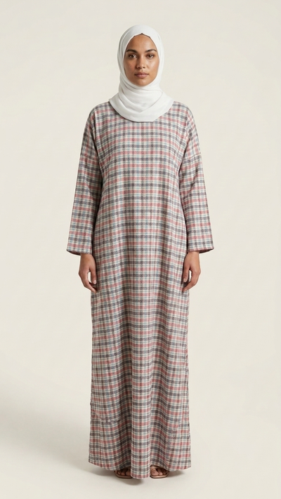 A woman wearing a long-sleeved, floor-length plaid abaya dress in a muted pink, red, and gray check pattern, paired with a white hijab, standing against a plain beige studio background.