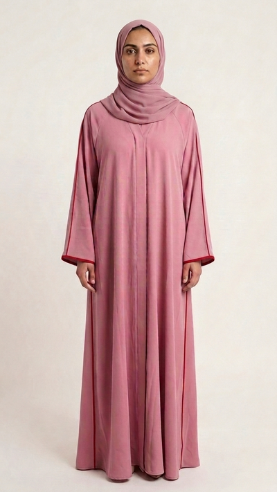 A woman wearing a long, flowing dusty pink abaya with a matching hijab, standing against a plain light background.