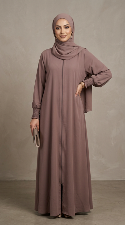 A woman modeling a long, modest abaya dress in a dusty mauve-brown color, paired with a matching hijab, standing against a textured gray studio backdrop.