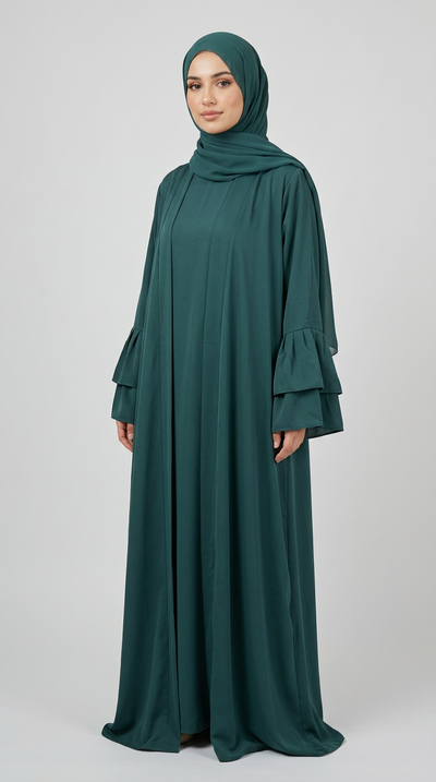 A woman wearing a dark teal green modest outfit consisting of a long flowing abaya with tiered ruffle sleeves and a matching hijab, standing against a light gray studio background.