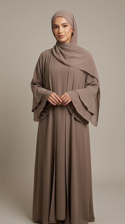A woman wearing a modest taupe-brown abaya with wide flared sleeves and a matching hijab, standing against a plain warm gray studio background.