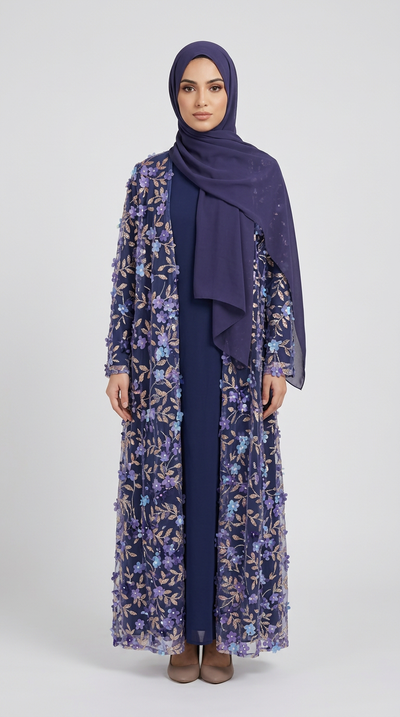 A woman modeling a long navy blue modest outfit with a matching hijab, featuring a sheer floral embroidered outer layer in purple, blue, and gold tones, photographed against a light gray studio background.