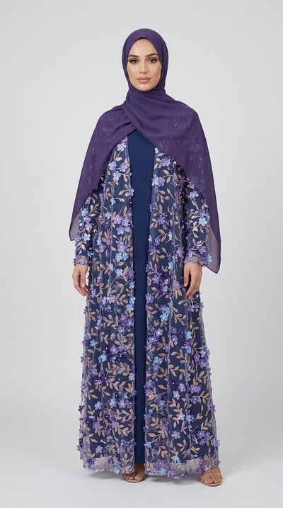A woman modeling a long navy blue floral embroidered abaya with a matching purple hijab, standing against a light gray studio background.