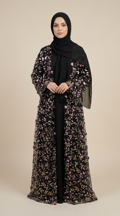 A woman wearing a long black abaya with dense floral embroidery and sequins in gold, pink, and purple tones, paired with a solid black hijab, standing against a solid beige studio background.
