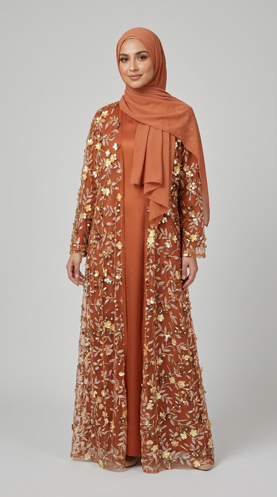 A woman modeling a rust-orange maxi abaya with gold floral embroidery and a matching draped hijab, photographed against a neutral light gray studio background.