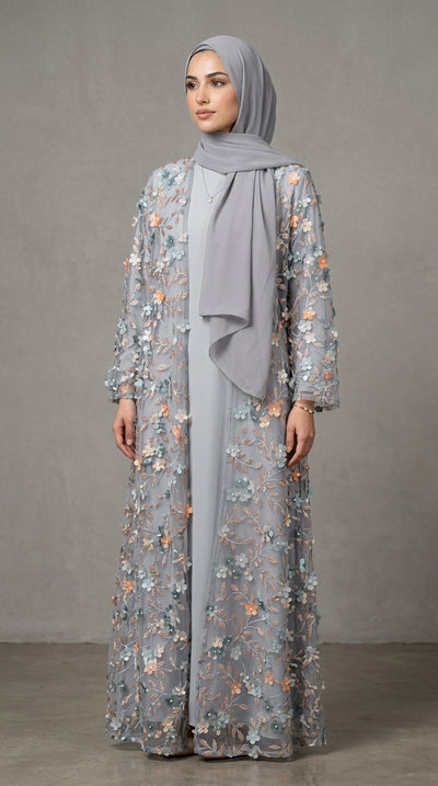 A woman wearing a long, light gray abaya dress with intricate multicolor floral embroidery and a matching draped hijab, standing against a neutral gray studio backdrop.