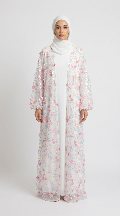 A woman wearing a long white abaya with a pink and multicolor floral patterned overlay jacket, paired with a white hijab, standing against a light gray studio background.