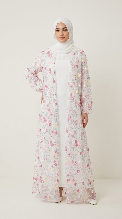 A woman wearing a long white abaya dress with a pink and multicolor floral print overlay, paired with a white hijab, standing against a plain light beige studio background.
