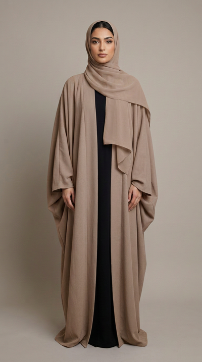 A woman modeling a long taupe abaya with a matching draped hijab over a black dress, photographed against a solid neutral studio background.