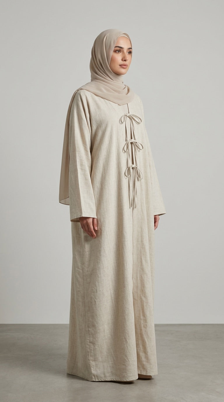 A woman wearing a long, loose beige abaya dress with a high neck and front tie details, paired with a matching beige hijab, standing against a neutral light gray studio background.