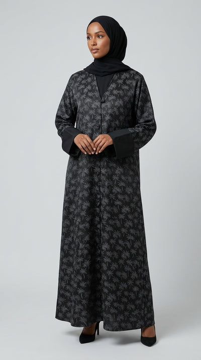 A woman wearing a long black abaya dress with a brown floral pattern, a black inner top, and a black hijab, standing against a light gray studio background.