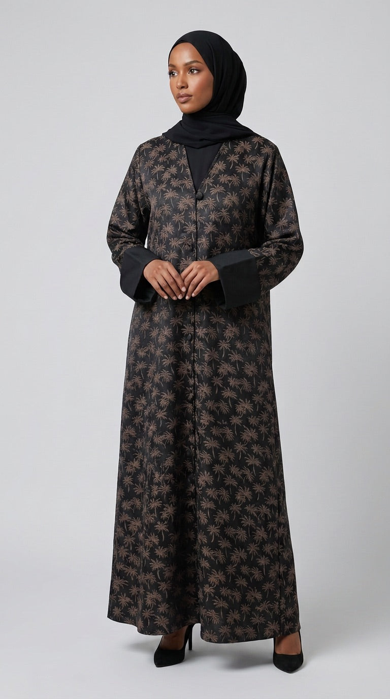 A woman wearing a long black abaya dress with a brown floral pattern, a black inner top, and a black hijab, standing against a light gray studio background.
