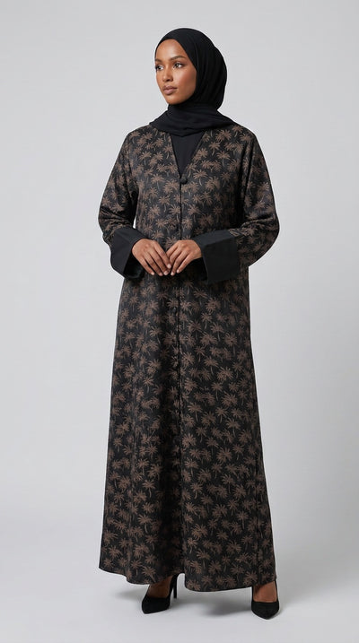 A woman wearing a long black abaya dress with a brown floral pattern, a black inner top, and a black hijab, standing against a light gray studio background.