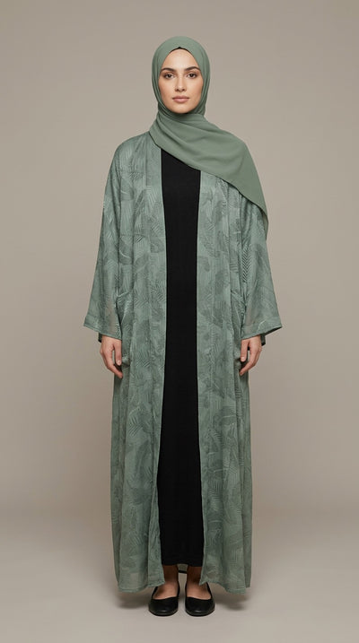 A woman modeling a long sage green abaya with a matching hijab, worn over a black dress, against a solid beige studio background.