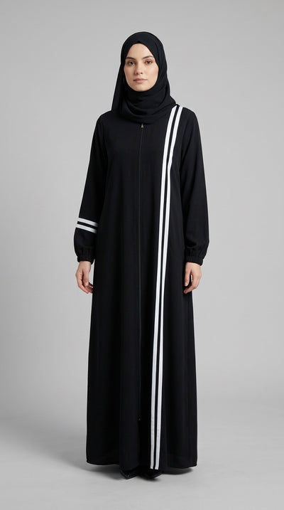 A woman wearing a long black abaya with white vertical stripes down the front and on the sleeves, paired with a black hijab, standing against a neutral gray studio background.