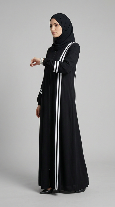 A woman wearing a long black abaya with white side stripes and a matching black hijab, standing against a plain light gray studio background.
